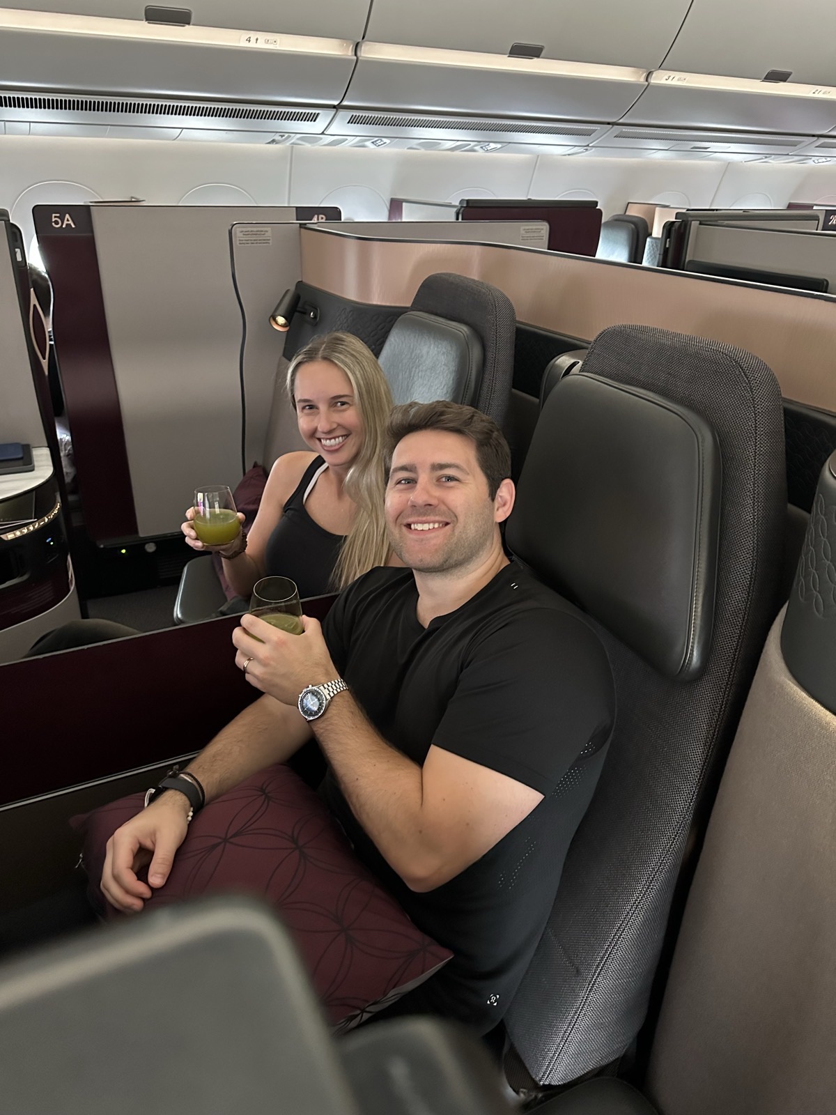 Mike and Christie smiling with drinks in their neighboring business-class seats, surrounded by private cabin partitions