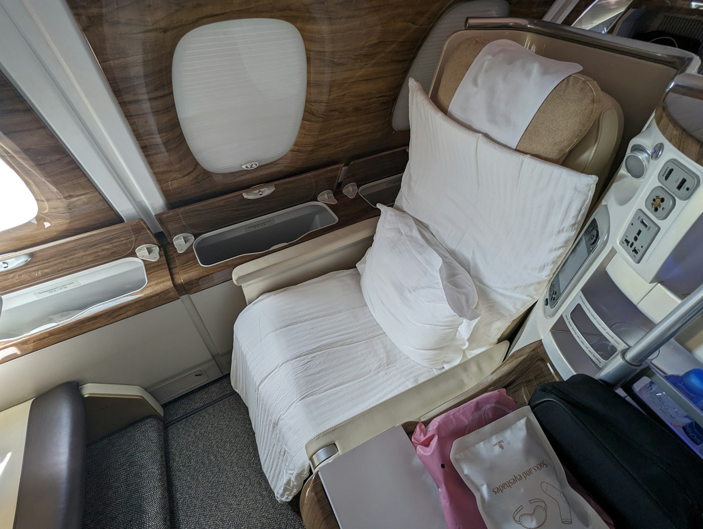 An empty Emirates business-class window seat prepared with a pillow and bedding, beside wood-finished cabin panels